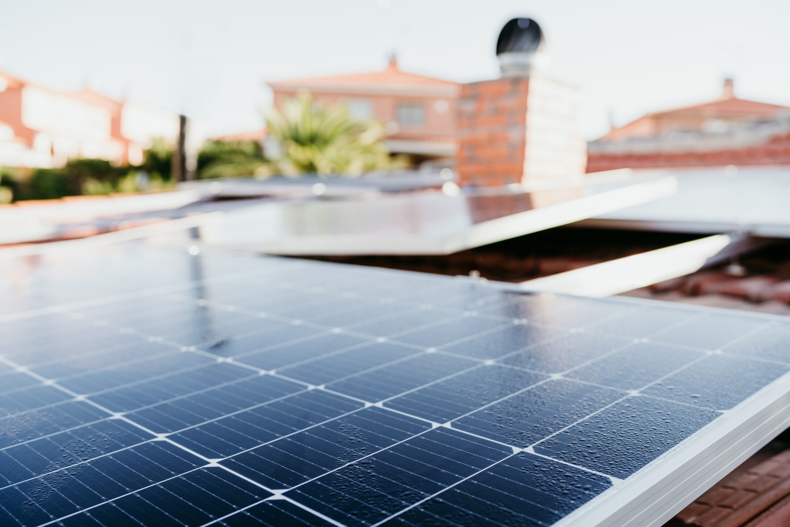 Close-up of solar panels installed on a rooftop, with houses and a brick chimney visible in the background.