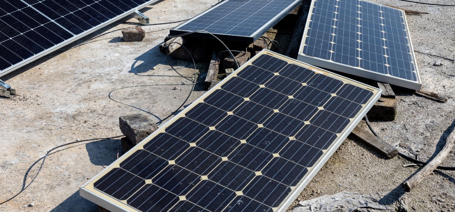 Three solar panels are positioned on a rough, concrete surface outdoors, with wires and wooden pieces scattered around&mdash;conditions that can contribute to common solar panel damage causes.