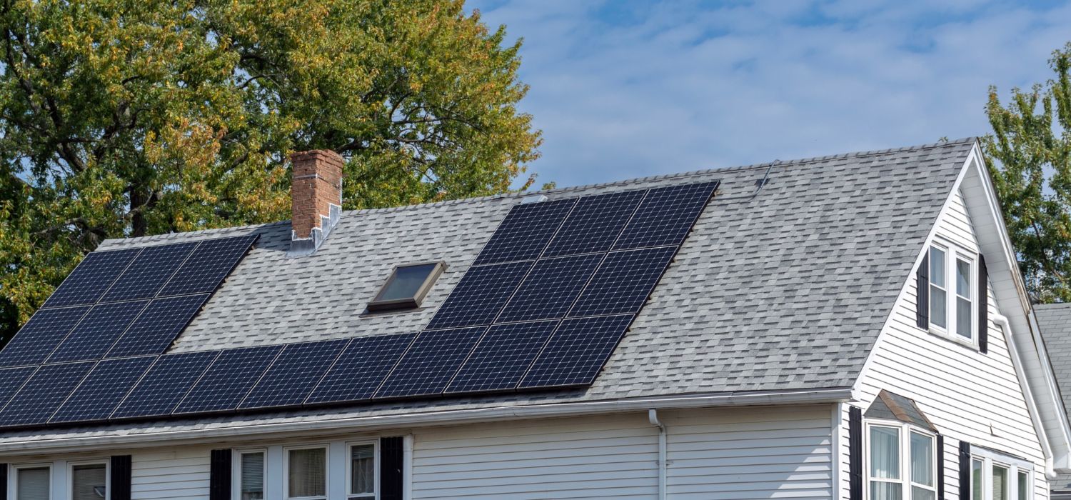 A white house with gray shingles features multiple solar panels on its roof, illustrating why solar energy is so important; trees and a blue sky complete the background.